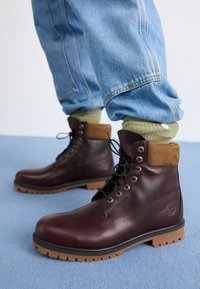 Stivali in pelle marrone con gambale in suede, punta rotonda, lacci neri e suola in gomma spessa. Indossati con jeans azzurri chiari e calzini verdi.