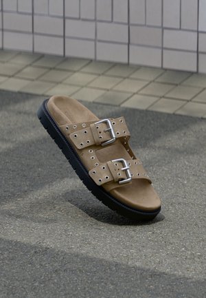 Sandale beige à enfiler avec semelle noire, deux brides réglables avec boucles argentées et plusieurs œillets, posée sur un sol texturé près d’un mur carrelé.