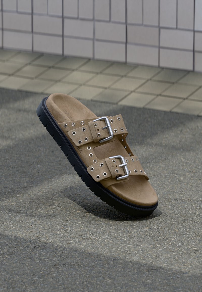 Ταμπά σανδάλι χωρίς λουράκι με μαύρη σόλα, δύο ρυθμιζόμενα λουριά με ασημένια αγκράφες και πολλαπλές οπές, τοποθετημένο σε υφασμένο έδαφος κοντά σε πλακιδωτό τοίχο.