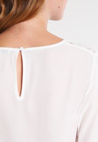 Blusa bianca con apertura a cuore sul retro, chiusa da un bottone. Tessuto liscio con delicati accenti in pizzo sulle spalle.