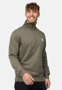 Oliven grøn zip-up sweater med høj krave, tekstureret stof, lange ærmer og et logo på brystet. Har en kontrasterende lynlås.