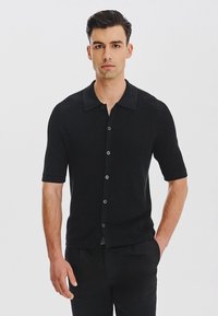 Mężczyzna stojący na białym tle, ubrany w czarną koszulę z krótkim rękawem z zapięciem na guziki oraz czarne spodnie, z jedną ręką w kieszeni.