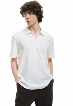 White short-sleeve polo shirt with a quarter zip, textured fabric, and a flat collar; worn with dark pants, hands in pockets.