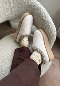 Pluizige crèmekleurige klompen met een tan-zool, geaccentueerd door bruine stiksels, gecombineerd met beige en goudgestreepte sokken op een gestructureerde stoel.
