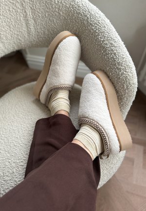 Pluizige crèmekleurige klompen met een tan-zool, geaccentueerd door bruine stiksels, gecombineerd met beige en goudgestreepte sokken op een gestructureerde stoel.