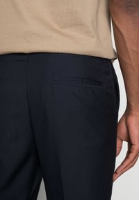 Tmavě modré kalhoty z hladké tkaniny, vybavené dvěma zadními kapsami s jemným lemováním, spárované s světle béžovou košilí.