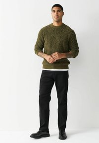 Olivovo zelený pletený sveter s textúrovaným vzorom, skombinovaný s čiernymi nohavicami a čiernymi mokasínami. Model stojí s rukami zloženými.