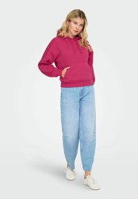 Femme avec de longs cheveux blonds portant un sweat à capuche magenta, un jean bleu clair ample et des baskets blanches, se tenant avec une main dans la poche de son sweat.
