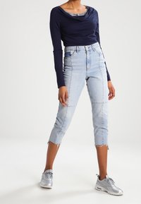 Navy-blaues Langarm-Oberteil mit Schalkragen, kombiniert mit hellblauen, verkürzten Jeans mit fransigen Säumen und silbernen Sportschuhen.