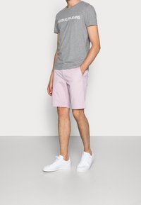 Camiseta de algodón gris con "CALVIN KLEIN JEANS" impreso en blanco, combinada con pantalones cortos rosa claro y zapatillas blancas. Conjunto casual con un corte relajado.