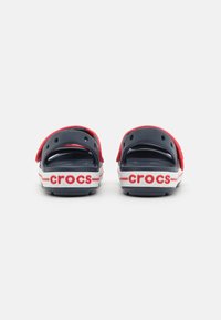 Punaiset ja laivastonsiniset Crocsit, joissa on avoimet kannat, ilmanvaihtoaukot sekä valkoinen "crocs"-logo, kontrastiväriset raidat ja teksturoitu pohjallinen.