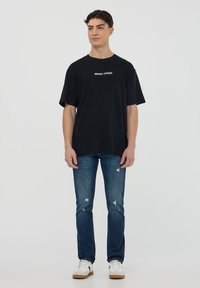 T-shirt nero a maniche corte con la scritta "WEST COAST" in bianco, abbinato a jeans blu strappati e sneakers bianche con accenti color sabbia.