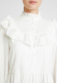 Vit långärmad klänning med volangkrage och bröst, med en texturerad tyg med ett subtilt rutmönster och knapprygg.