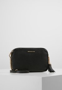 Černá kožená crossbody taška Michael Kors se zlatým řetízkem a detailem střapce, umístěná na bílé ploše na šedém pozadí.