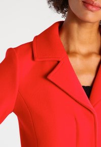 Rode wollen blazer met een geknipte kraag, gestructureerde schouder en strakke lijnen. De stof heeft een gladde textuur met een op maat gemaakte pasvorm.