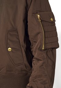 Brown bomber jacket featuring ribbed cuffs, a zippered sleeve pocket, and gold hardware accents. The fabric appears smooth with a matte finish.