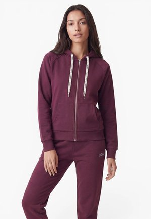 Jeune femme portant un sweat à capuche bordeaux zippé avec des cordons blancs et un pantalon de jogging bordeaux assorti, debout devant un fond blanc uni.