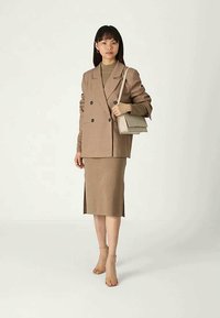 Blazer beige à double boutonnage, jupe beige ajustée et sandales à talons en cuir tan. Le blazer est orné de grands boutons ; la jupe présente une fente sur le côté.