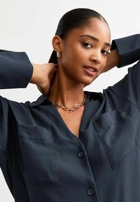 Camicia blu navy con bottoni in tessuto setoso, dotata di due tasche sul petto, abbinata a una collana in oro dallo stile massiccio e orecchini a cerchio dorati.