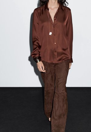 Femme portant une chemise ample en soie marron à boutons et un pantalon en daim marron, accessoirisée avec des bracelets et un long collier avec un pendentif.