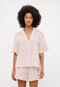 Ensemble de pyjama blanc à manches courtes avec des motifs de cœurs rouges. Le haut présente un plastron à boutons et une coupe décontractée ; le short a un ourlet volé.