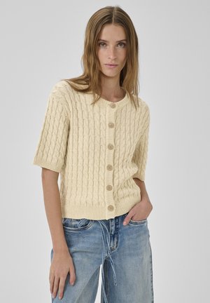 Frau trägt eine cremefarbene, kurzärmelige Strickjacke mit Zopfmuster und Knöpfen sowie lockere hellblaue Jeans, posiert mit einer Hand in der Tasche.