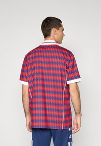 Czerwono-niebieska koszulka polo z geometrycznym wzorem, z krótkimi rękawami, białym kołnierzykiem i kontrastującymi detalami. Wykonana z lekkiego materiału.