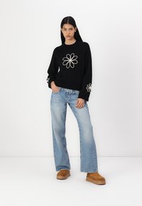 Svart genser med hvit blomster broderi; lys blå vide jeans; brune chunky plattformsko; modellen står med hender i lommene.