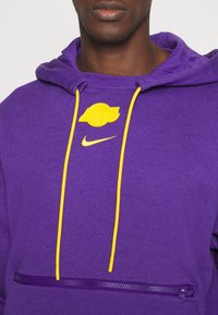 Sweat-shirt violet à capuche, accents jaunes, et poche zippée à l'avant. Présente le logo Nike et un graphisme des Lakers sur la poitrine.