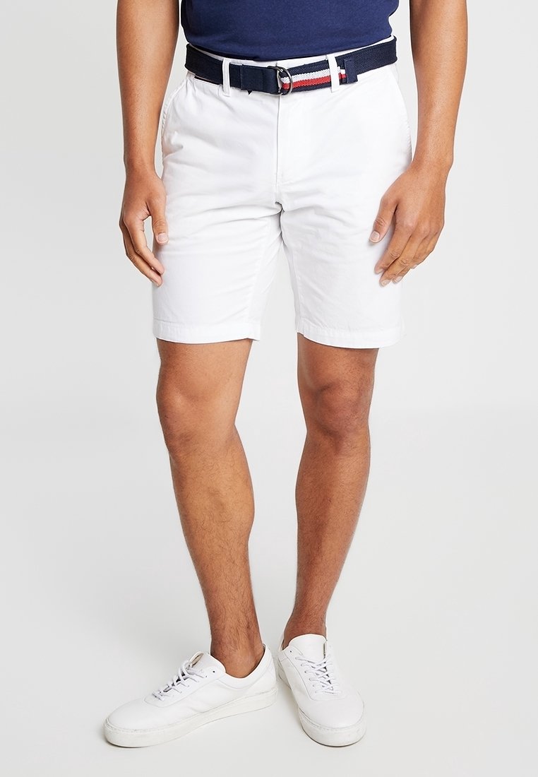Homme portant un short blanc, une ceinture bleu marine avec des rayures rouges et blanches, des baskets blanches et une chemise bleu marine, debout avec les mains détendues sur les côtés.