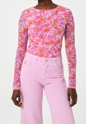 Femme portant un haut à manches longues à motif floral rose et un pantalon taille haute rose clair, debout devant un fond uni.
