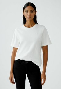 Weißes T-Shirt mit kurzen Ärmeln und rundem Halsausschnitt, aus weichem Material; getragen mit schwarzen Jeans, dezent mit einem kleinen Logo auf der Brust.