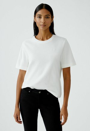 White short-sleeve t-shirt with a round neckline, made of soft material; worn with black jeans, subtly featuring a small logo on the chest.
