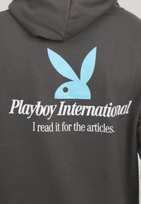 Sudadera gris oscuro con un logo de Playboy en azul claro y texto blanco que dice "Playboy International, la leo por los artículos."