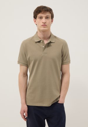 Young man with short brown hair wearing a short-sleeve olive green polo shirt and dark pants, standing with one hand in pocket.