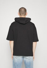 Schwarzer Kapuzenpullover mit kurzen Ärmeln, weicher Textur und lässiger Passform, kombiniert mit hellblauen, abgenutzten Jeans.