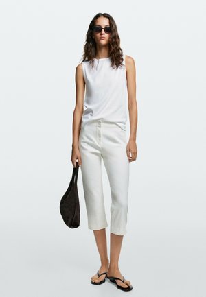 Vrouw met golvend haar die zwarte zonnebril draagt, een mouwloos wit top, witte cropped broek, zwarte sandalen en een zwarte tas bij zich heeft.