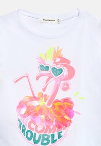 Ροζ φλαμίγκο με γυαλιά ηλίου σε σχ ήμα καρδιάς με γκλίτερ και πολύχρωμο κολιέ από φτερά, με το κείμενο «Εδώ έρχεται η φασαρία» σε λευκό πουκάμισο.