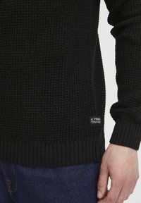 Suéter negro de punto con superficie texturizada y puños acanalados. Presenta una pequeña etiqueta en el lateral. Usado sobre jeans azul oscuro.