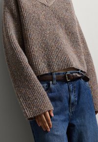 Maglione lavorato a maglia in beige tenue con una texture a costine e scollo a V. Abbinato a jeans blu a vita alta e una cintura in pelle marrone.