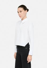 Camicia bianca a maniche lunghe con colletto a punta, decorata con dettagli perline sul colletto e sui polsini, abbinata a pantaloni neri.