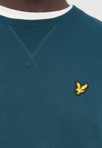 Mätová mikina s okrúhlym výstrihom, textúrovaným materiálom a žltým vyšívaným logom orla na ľavej strane hrude. Kontrastný biely golier.