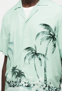 Άνδρας που φοράει ανοιχτοπράσινο πουκάμισο με κοντά μανίκια με μαύρα σχέδια από φοίνικες και λευκά λουλούδια στο μπροστινό μέρος.