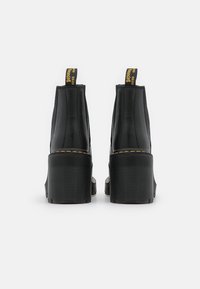 Mustat nahkaiset nilkkurit, joissa on paksu uritettu pohja, keltainen brändäys vetoketjussa ja sileä, kiiltävä pinta.