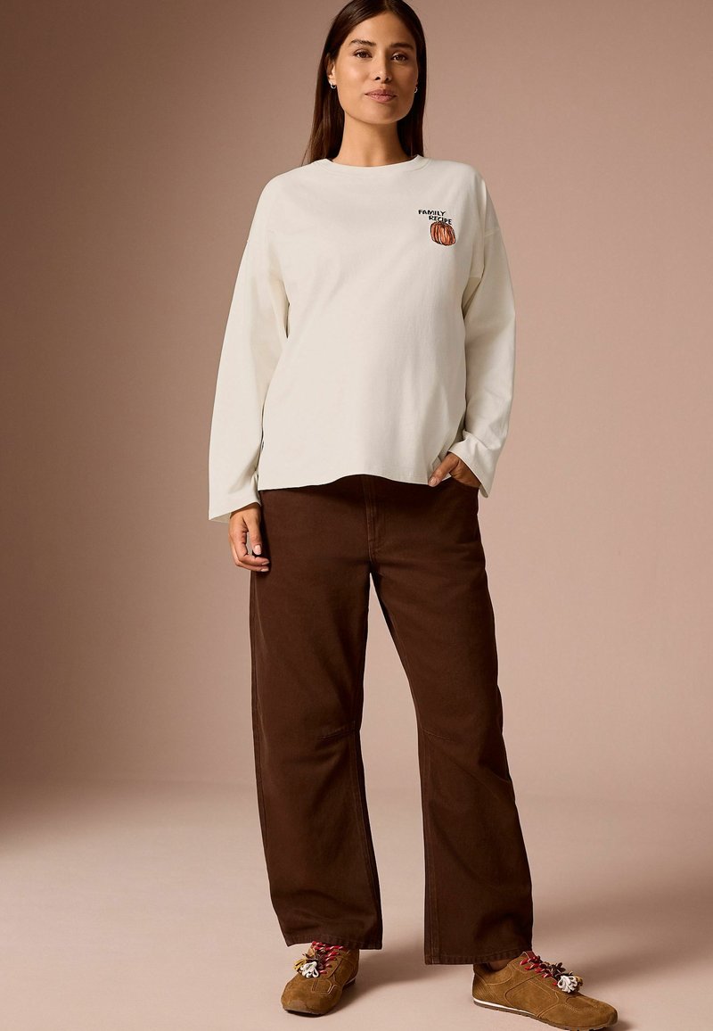 Woman wearing cream long-sleeve shirt with "Family Recipe" and pumpkin graphic, brown wide-leg pants, and brown lace-up shoes.