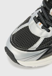 Basket de course en mesh noir avec des superpositions synthétiques argentées, tige texturée, lacets noirs et semelle intermédiaire blanche. Design angulaire avec accent orange.