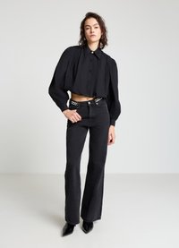 Schwarzes, kurz geschnittenes Hemd mit Knöpfen und aufgepufften Ärmeln, kombiniert mit hoch taillierten, schwarzen, ausgestellten Jeans und spitzen schwarzen Absätzen.