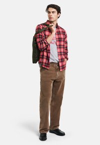 Brązowe spodnie z korduroju w połączeniu z czerwoną i czarną koszulą w kratę na szarym t-shircie z nadrukiem. Czarne buty i oliwkowy plecak.