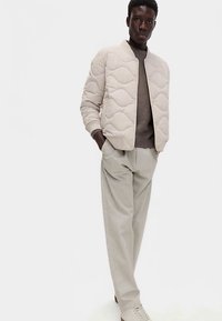 Gewatteerde beige bomberjack met een gestructureerd golfpatroon, gecombineerd met een bruine trui, lichtbeige broek en lichte sneakers.