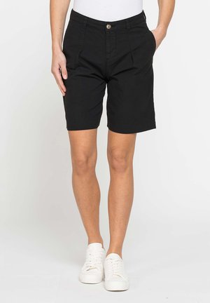 Persona che indossa pantaloncini neri fino al ginocchio, scarpe da ginnastica bianche e una maglietta bianca con una mano in tasca, su uno sfondo semplice.